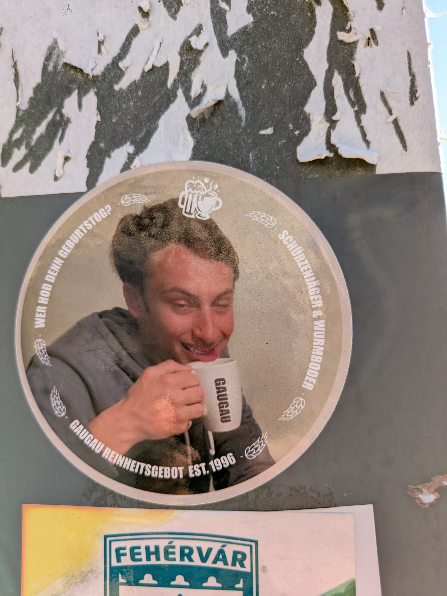 This is a round sticker on a wall. It features a man drinking from a mug with the word "GAUGAU" on it. The sticker has text around the edge, including "WER HOD DENN GEBURTSTOG?", "SCHURZENJAGER & WURMBOODER" and "GAUGAU REINHEITSGEBOT EST. 1996".