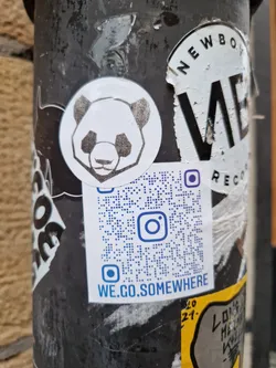 A round sticker featuring a stylized panda design. Below it is a QR code with the text 'WE.GO.SOMEWHERE' and an Instagram logo. The sticker is affixed to a dark-colored pole with other stickers.