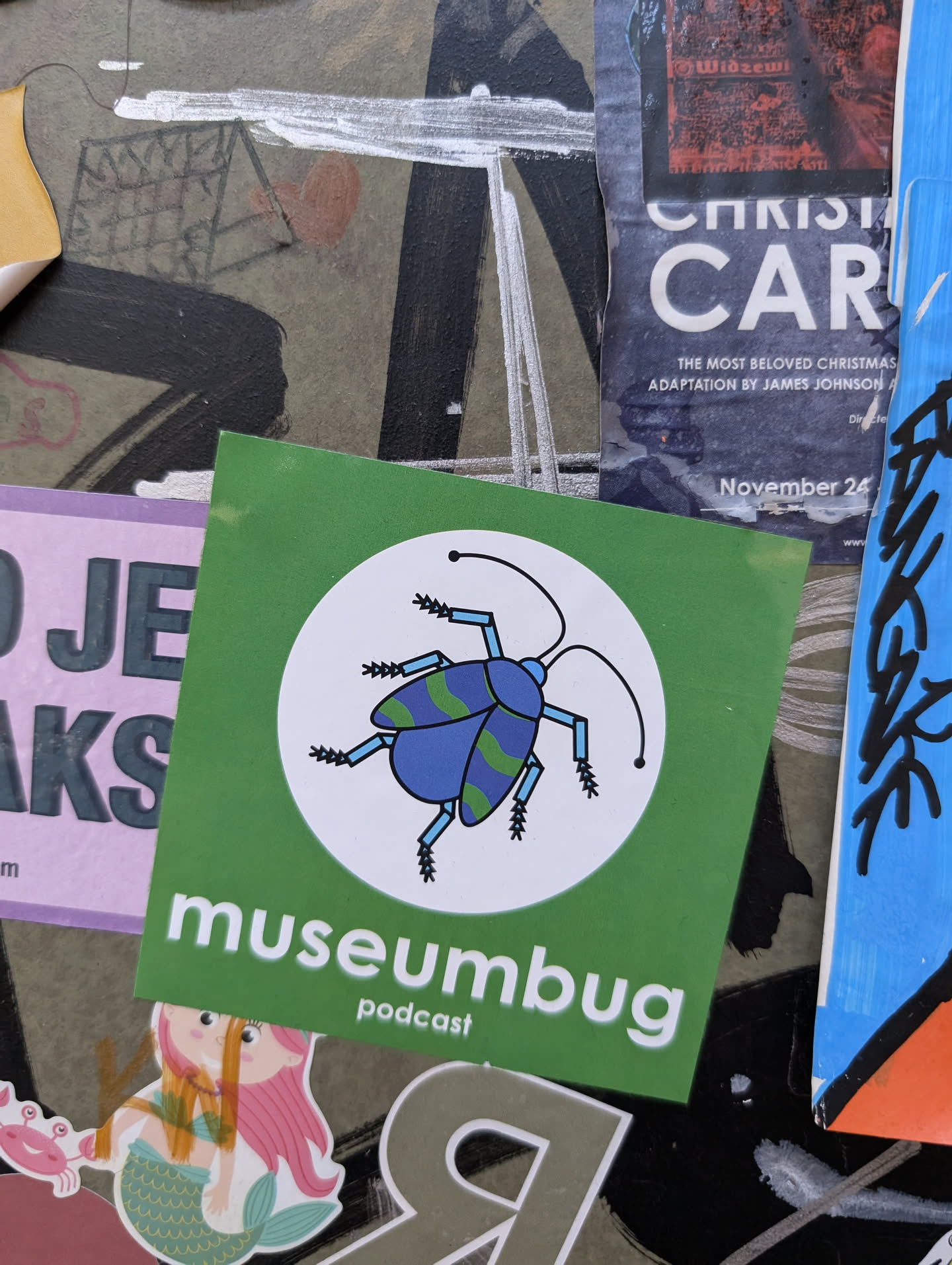 A green square sticker with a white circle in the center featuring a blue insect with green stripes. The text "museumbug podcast" is written below the circle.