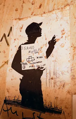 Black and white stencil artwork depicting a person holding a sign that reads: I SAVED MYSELF TODAY.  The artwork is distressed, showing signs of wear and tear. There is additional text on the sign including a phone number and website address.