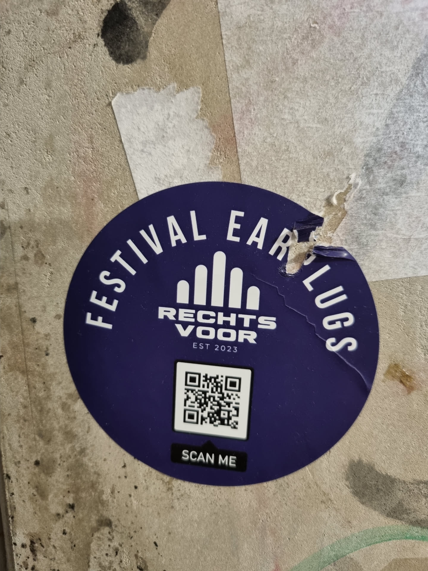 Purple round sticker with the text "Festival Earplugs" written in a circle around a logo and a QR code. Below the QR code it says "Scan me". The logo shows five stylized mountains with the text "Rechts voor" and "Est 2023" underneath.