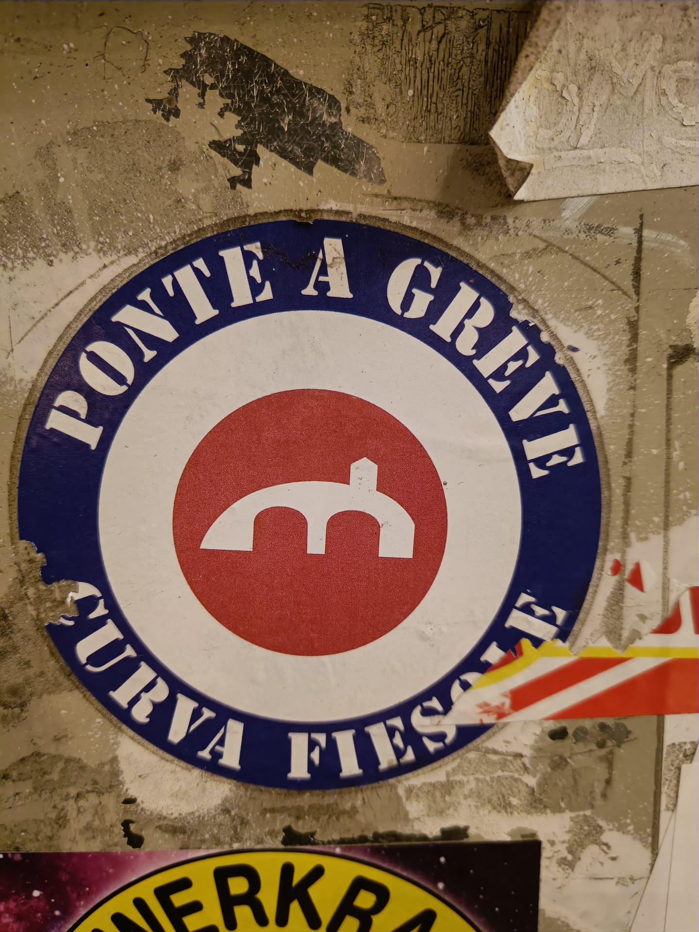 A round sticker with the text "PONTE A GREVE CURVA FIESOLE" in a blue circle, and a red circle in the center with a symbol that looks like a bridge. The sticker is a bit worn and is stuck on a wall with other stickers.