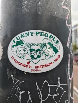 A vintage oval sticker featuring three cartoon faces with the text "FUNNY-PEOPLE",  "TT VASUMWEG 4C AMSTERDAM - NOORD (NDSM)" . The sticker is affixed to a dark-colored pole.