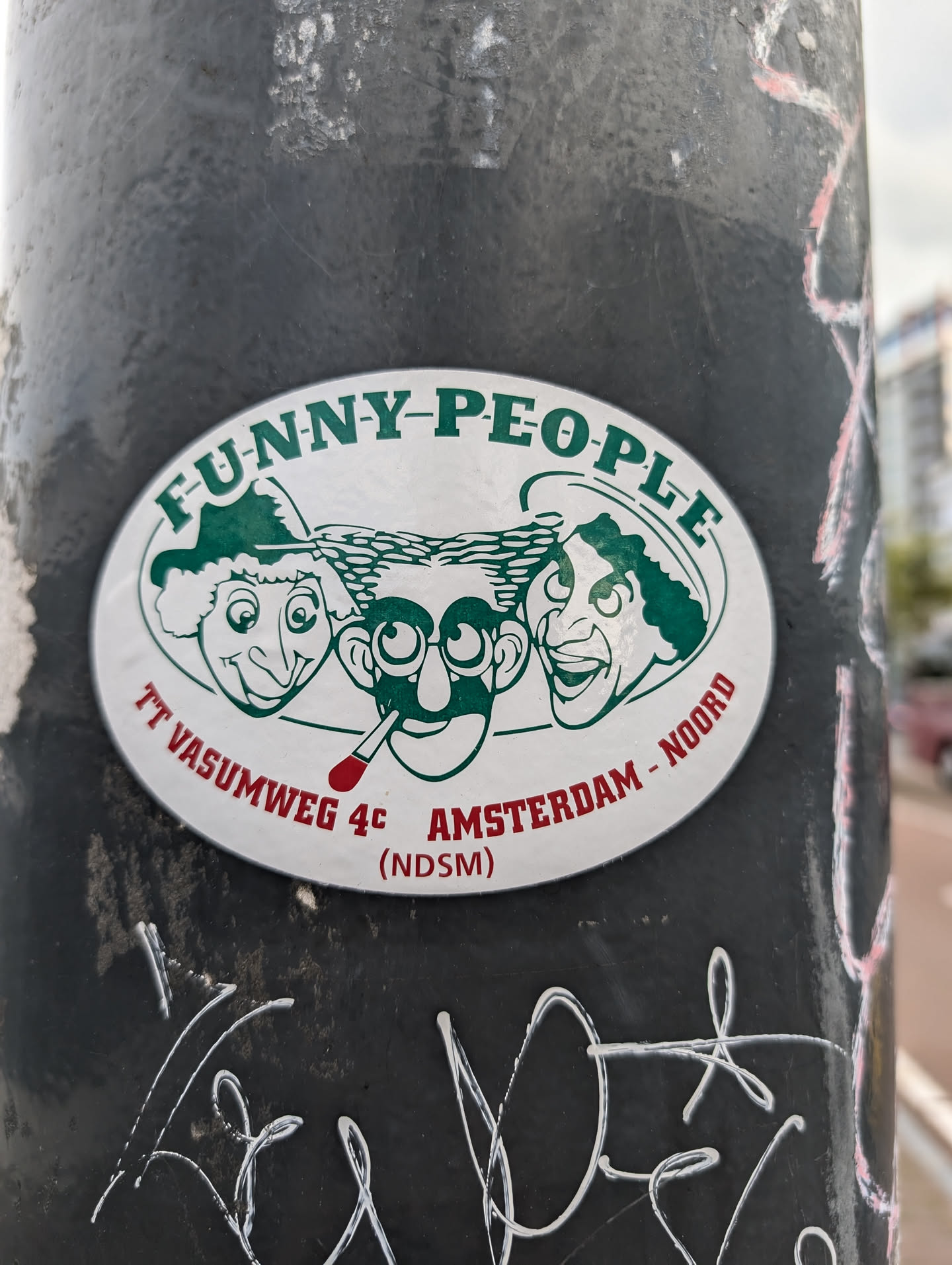 A vintage oval sticker featuring three cartoon faces with the text "FUNNY-PEOPLE",  "TT VASUMWEG 4C AMSTERDAM - NOORD (NDSM)" . The sticker is affixed to a dark-colored pole.