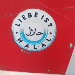 A round sticker with the word Halal written in Arabic and German. The sticker is white with a light blue ring around the Halal text. The text also says "Liebe ist Halal" which translates to "Love is Halal". There is a website address at the bottom of the sticker: www.liebeist-halal.de