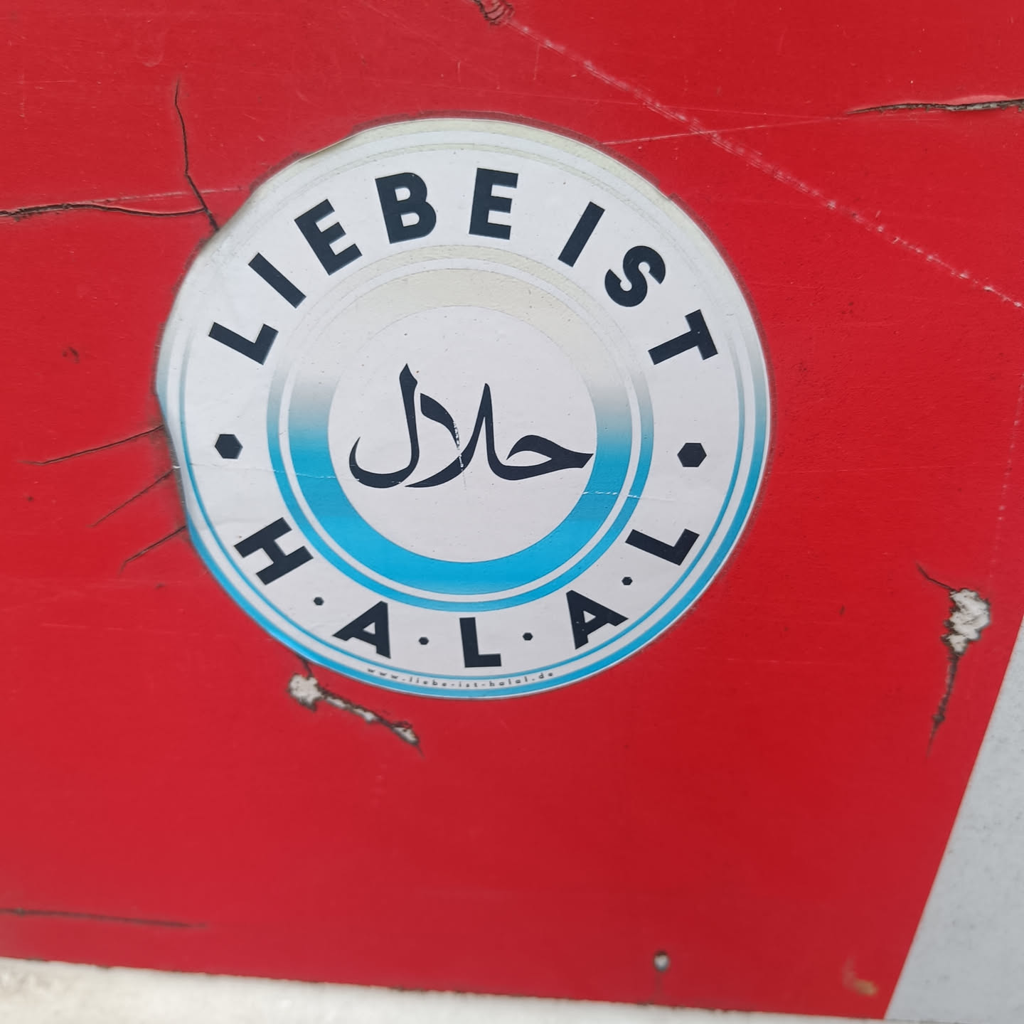 A round sticker with the word Halal written in Arabic and German. The sticker is white with a light blue ring around the Halal text. The text also says "Liebe ist Halal" which translates to "Love is Halal". There is a website address at the bottom of the sticker: www.liebeist-halal.de