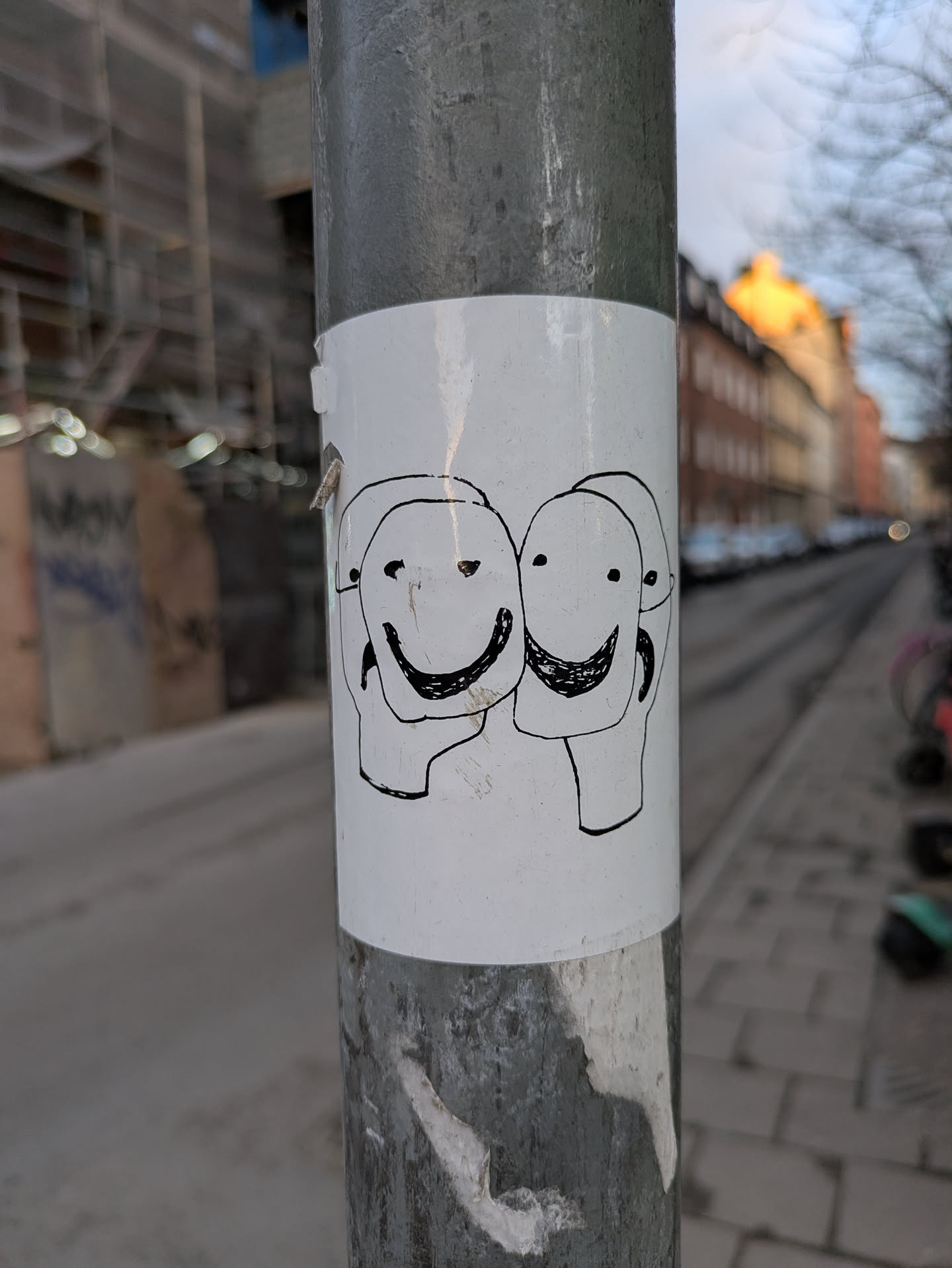 A sticker featuring a simple drawing of three heads with smiling faces. The drawing is in black ink on a white background. The sticker is affixed to a metal pole on a city street.