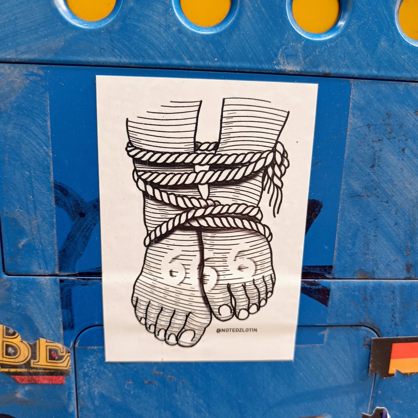 A black and white illustration of two feet bound by rope.  The style is reminiscent of old anatomical drawings. The sticker is affixed to a blue surface. There is an Instagram handle at the bottom.