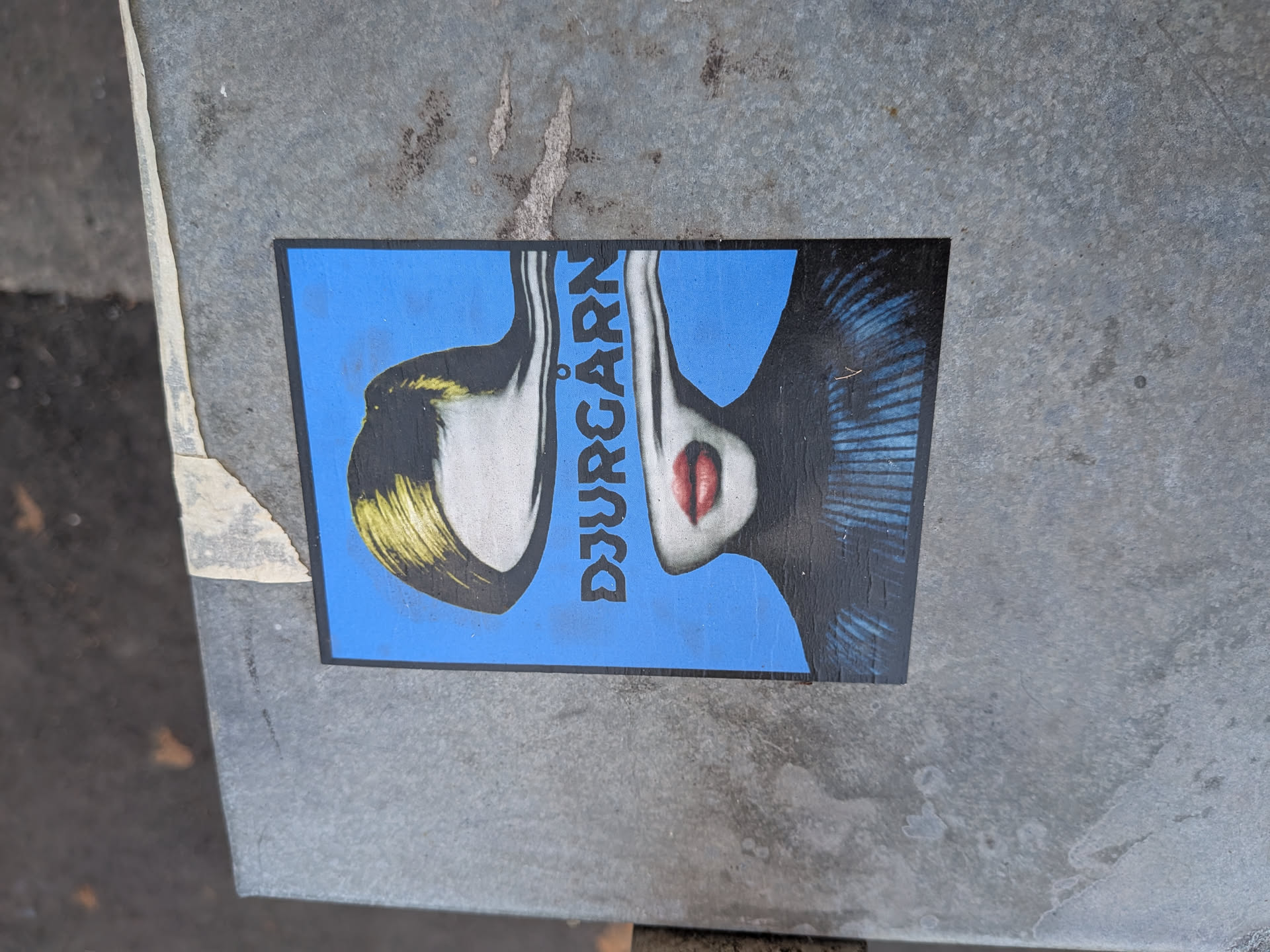 A rectangular sticker depicting a woman's face split vertically. The left side features blonde hair, while the right side shows red lips. The vertical text 'DJURGÅRN' is positioned between the two halves against a bright blue background.