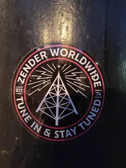Black and pink circular sticker with a radio tower graphic and text. The text includes 'Zender Worldwide', 'Est. 74', and 'Tune In & Stay Tuned'.