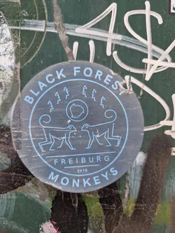 Round sticker with dark-gray background and light-blue text. It shows two monkeys facing each other, with trees and a circular symbol between them. The text around the image says: "Black Forest Monkeys" and "Freiburg 2018" in the middle.