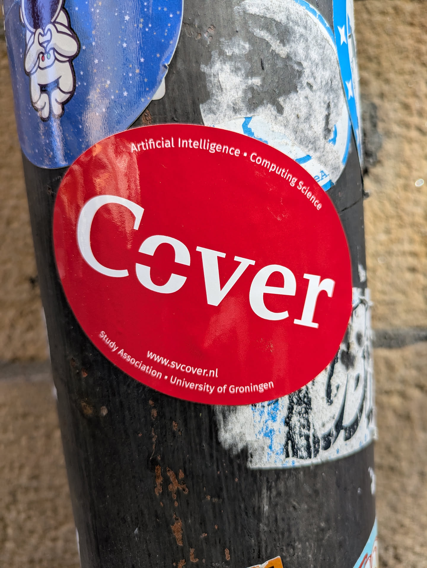 A round, red sticker on a pole featuring the word "Cover" in large, white, bold letters. Above the word "Cover" are the words "Artificial Intelligence" and "Computing Science" in a smaller font. Below the word "Cover" are the words "Study Association • University of Groningen" and the website "www.svcover.nl".