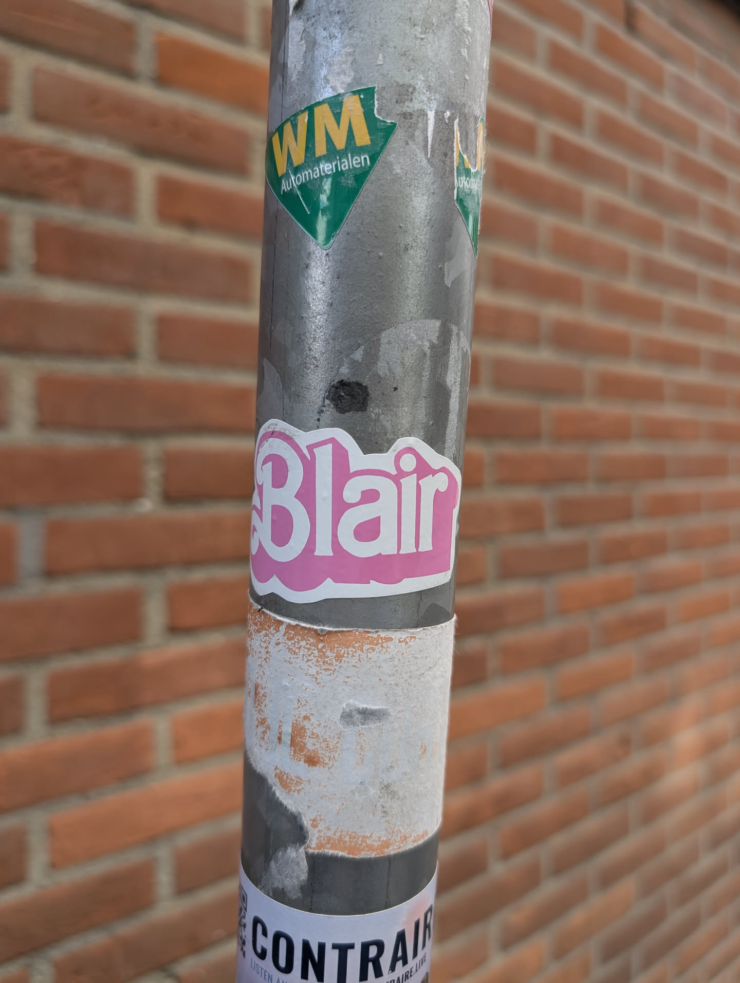 A pink sticker with the name 'Blair' in a playful font is adhered to a gray pole, which also bears several other stickers. The background features a brick wall, providing a common urban setting.