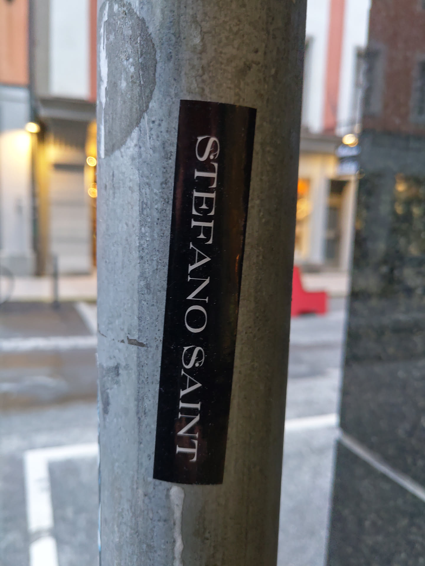 A black sticker with the text "Stefano Saint" written vertically in a minimalist, elegant font.  It's affixed to a weathered metal pole. 