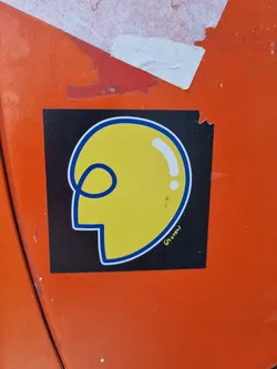 This sticker features a minimalistic design of a yellow head with simple blue lines. It has a black background and the artist's signature is visible at the bottom right corner.