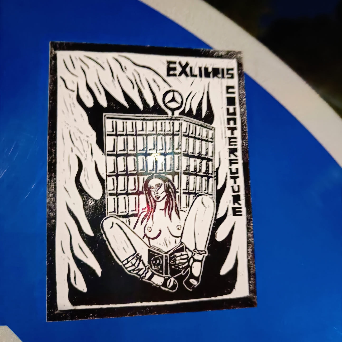 Black and white sticker depicting a topless woman sitting with a book in front of a burning building. The building has a Mercedes-Benz logo on top. The text "Ex libris counterculture" is written vertically on the sides.