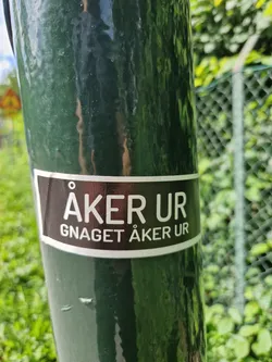 A black and white sticker with the text "ÅKER UR" in a larger font and "GNAGET ÅKER UR" in a smaller font underneath. It's attached to a dark green pole outdoors.