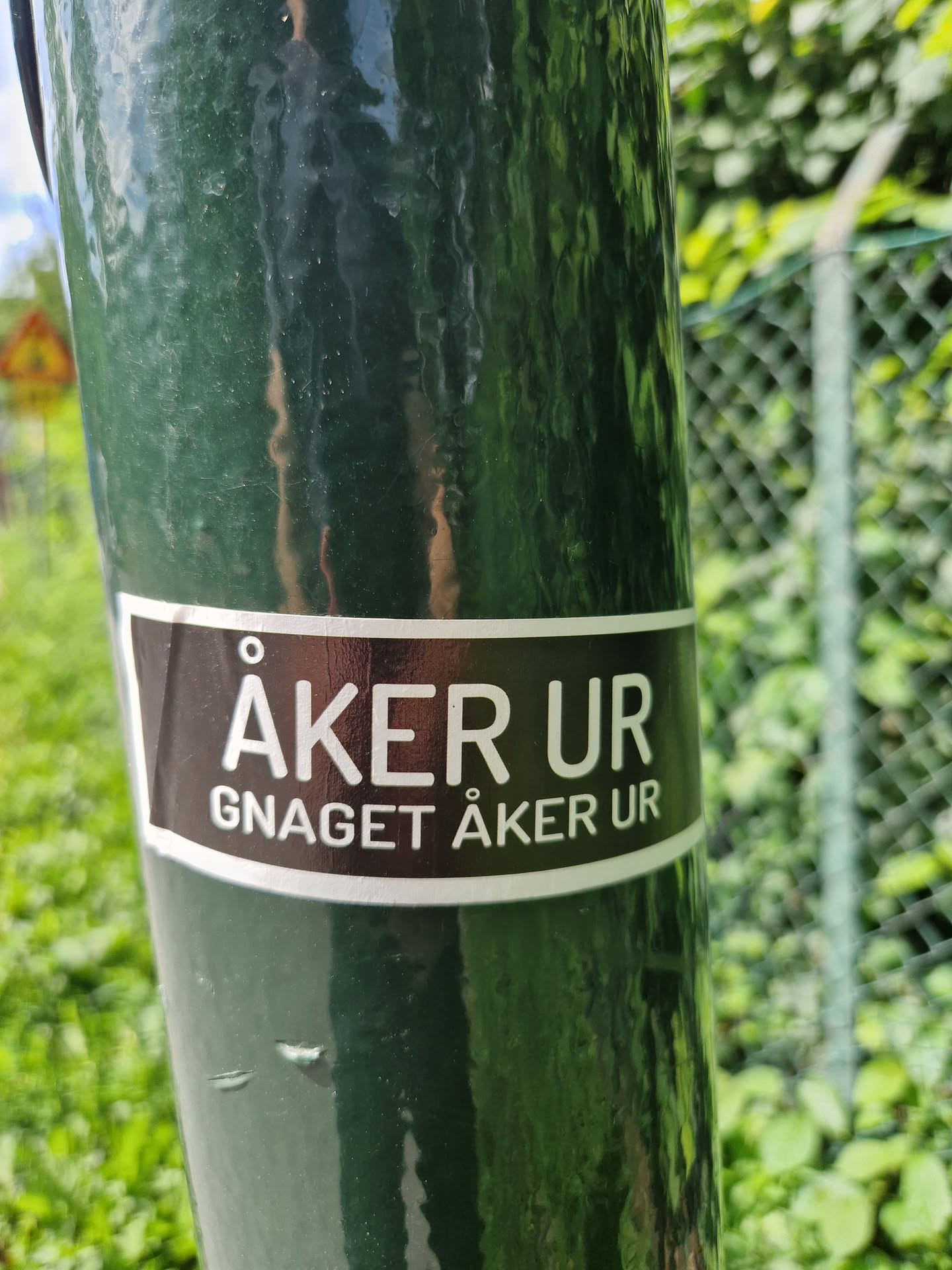 A black and white sticker with the text "ÅKER UR" in a larger font and "GNAGET ÅKER UR" in a smaller font underneath. It's attached to a dark green pole outdoors.