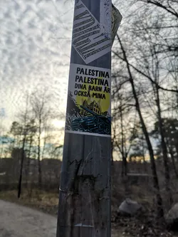 The sticker is a rectangular piece adhered to a metal pole. It features a stark graphic design with a primary color palette of black, yellow, and blue. The upper portion displays bold, black uppercase text on a white background. Below the text, a large, bright yellow circle, possibly representing a sun or moon, dominates the background. Overlaid on this yellow circle and extending downwards is a dark, silhouetted landscape of what appears to be destroyed or war-torn buildings. In the foreground, a detailed illustration of a blue military tank is prominently featured, moving across the lower part of the scene. The overall aesthetic is impactful and conveys a strong political or social message.