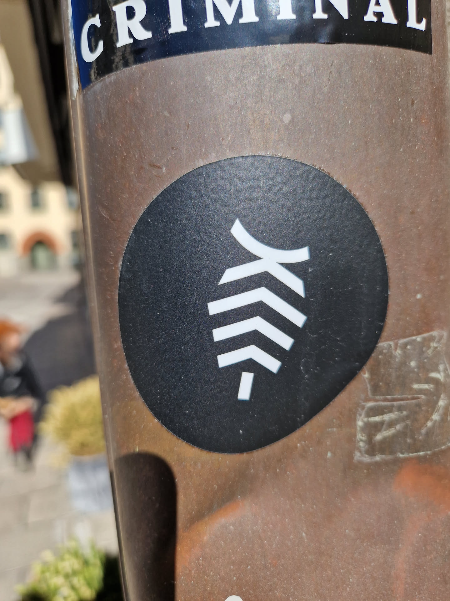 A black and white circular sticker with a stylized leaf or feather design. The sticker is affixed to a brown metal pole. 