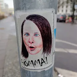 A sticker depicting a woman with a surprised expression.  The word "Hömma!" is written below the image in a stylized font. The style is reminiscent of hand-drawn illustration or sketch.