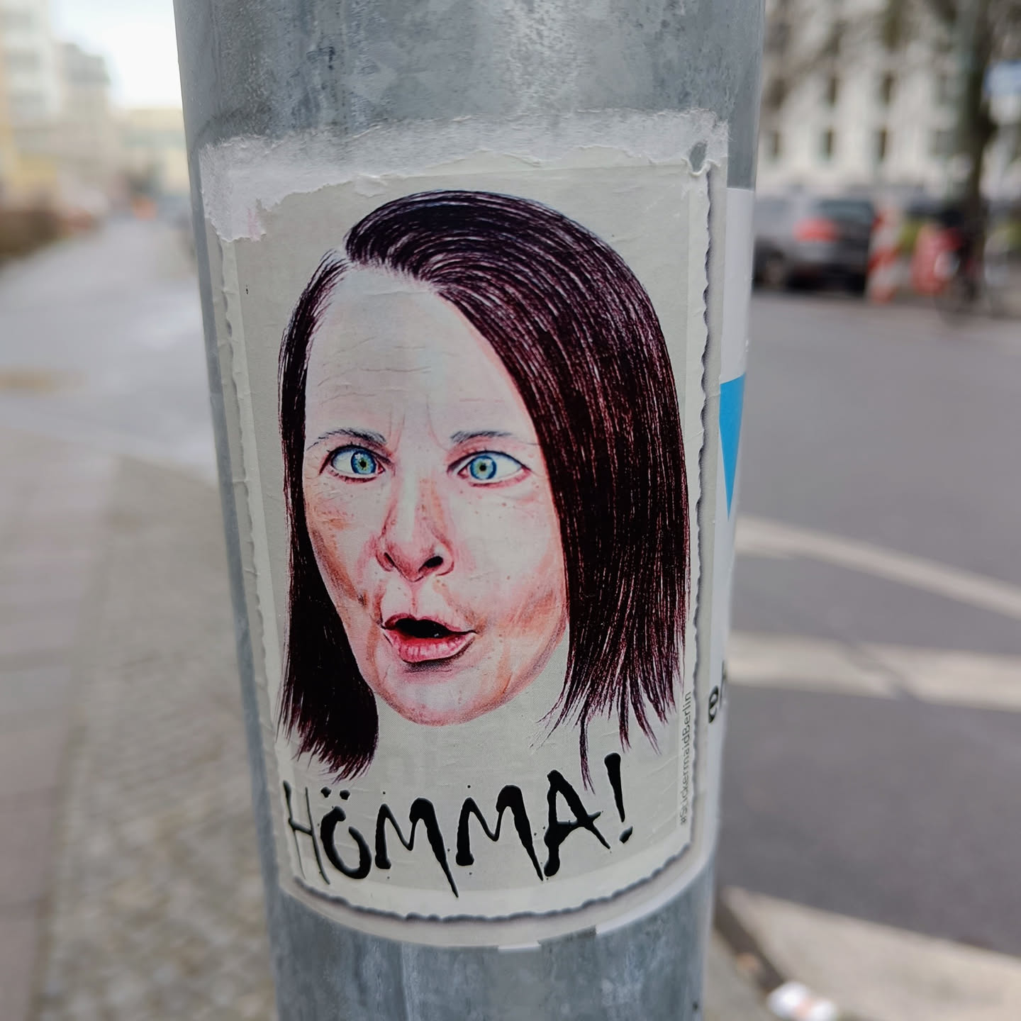 A sticker depicting a woman with a surprised expression.  The word "Hömma!" is written below the image in a stylized font. The style is reminiscent of hand-drawn illustration or sketch.