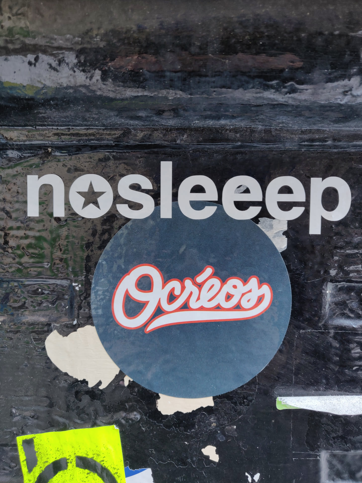 Round sticker with the text "Ocréos" in a stylized red and white font on a dark blue background.  The sticker is placed on a dark surface with other stickers and markings.  Above the sticker is a banner with the text "nosleep" in a sans-serif font.