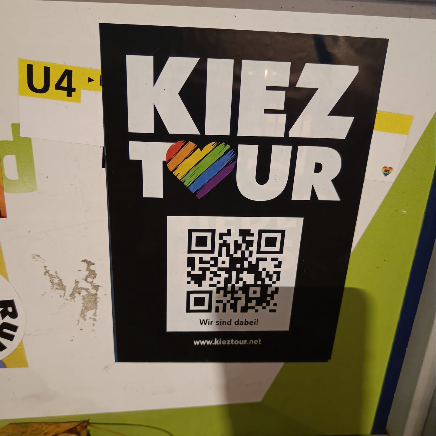 A black and white sticker with a rainbow heart in the middle and a QR code at the bottom.  The text on the sticker says "Kiez Tour", "Wir sind dabei!" and "www.kieztour.net".