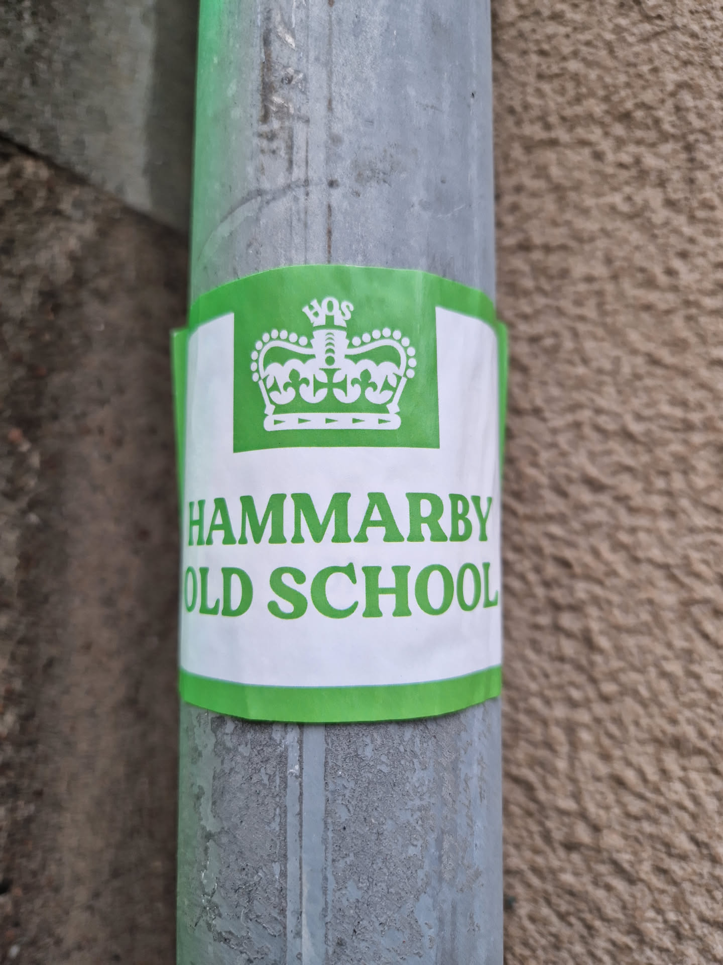 A green and white sticker with the text "HAMMARBY OLD SCHOOL" and a crown logo is attached to a metal pipe. The sticker appears to be weathered and has been exposed to the elements.
