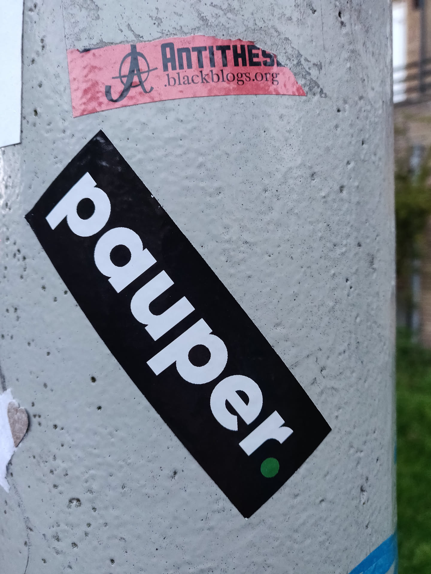 A black and white sticker with the word "pauper" written in a bold, sans-serif font. There's a small green dot at the bottom right corner of the sticker.