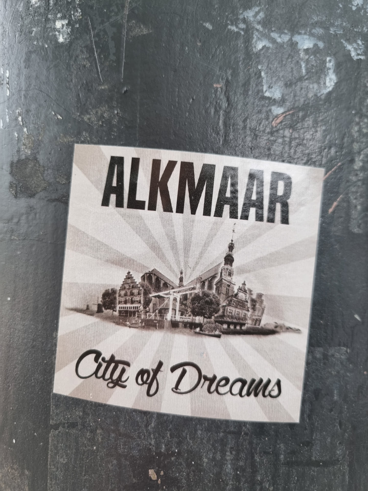 A vintage-style sticker depicting Alkmaar, Netherlands. The sticker features a sepia-toned image of the city's iconic waterfront and buildings, with the city's name prominently displayed.  The tagline 'City of Dreams' adds a whimsical touch.