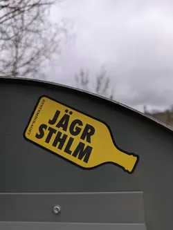 A yellow sticker shaped like a bottle with the text "Jägr Sthlm" on it, adhered to a gray metal surface. The sticker is slightly weathered, and the background includes a bare tree and overcast sky.