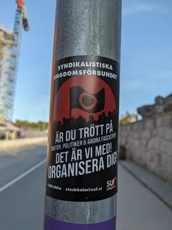 A sticker from the Syndikalistiska Ungdomsförbundet (Syndicalist Youth League) expressing frustration with corruption, politicians, and fascists, and encouraging organization.  The sticker features a black flag with a red circle and features their contact information.