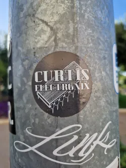 A round sticker with the text 'Curtis Electronix' and an image of an integrated circuit. The sticker is affixed to a metal pole.