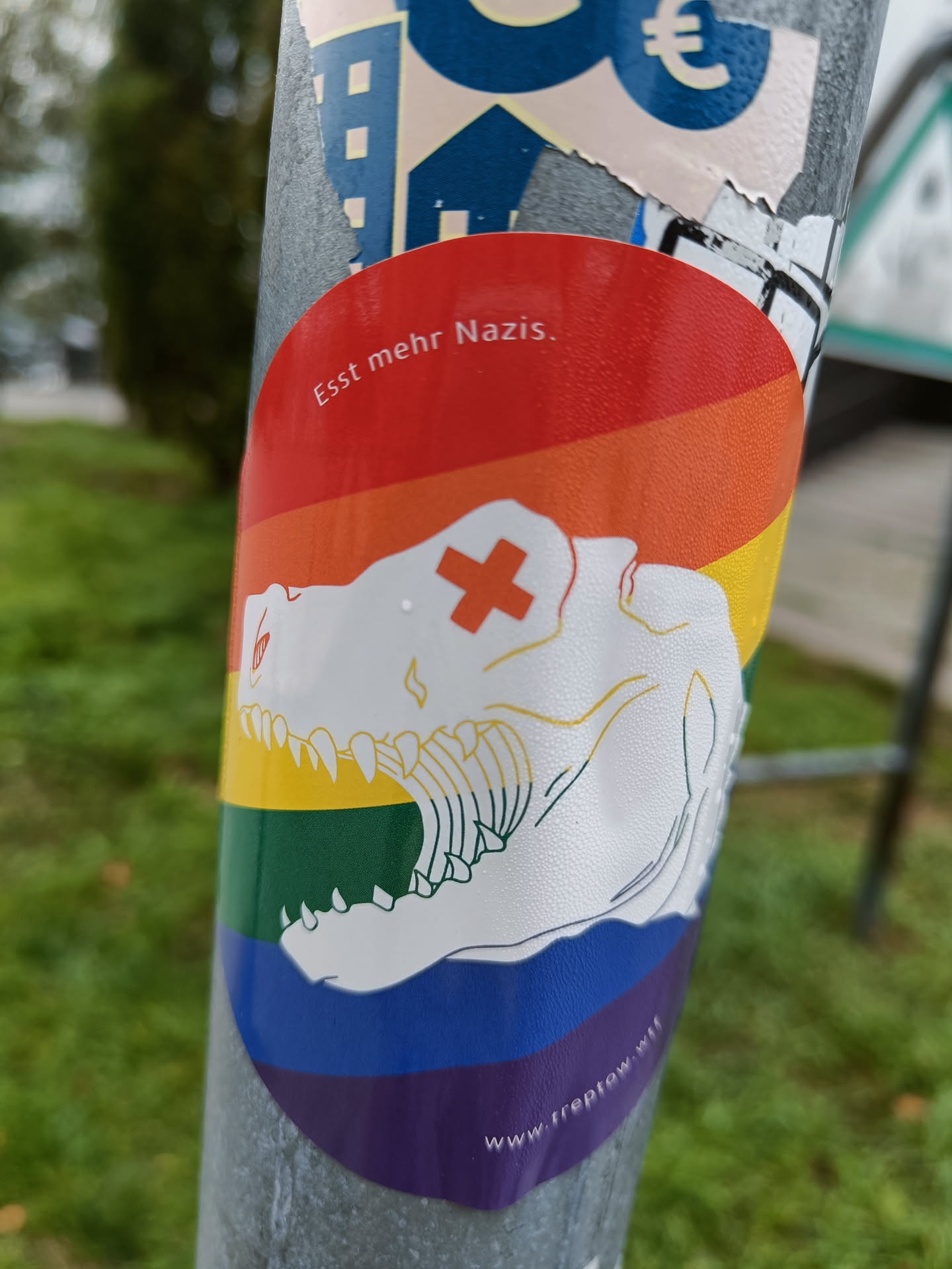A round sticker with a rainbow colored background. On the sticker is a drawing of a T-Rex skull with a cross on its forehead. At the bottom of the sticker there is a website address: www.freeplow.at