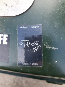A black sticker with white text. The sticker has the artist's name oTrOgEn and social media links. The text on the sticker is hand-written and slightly messy.