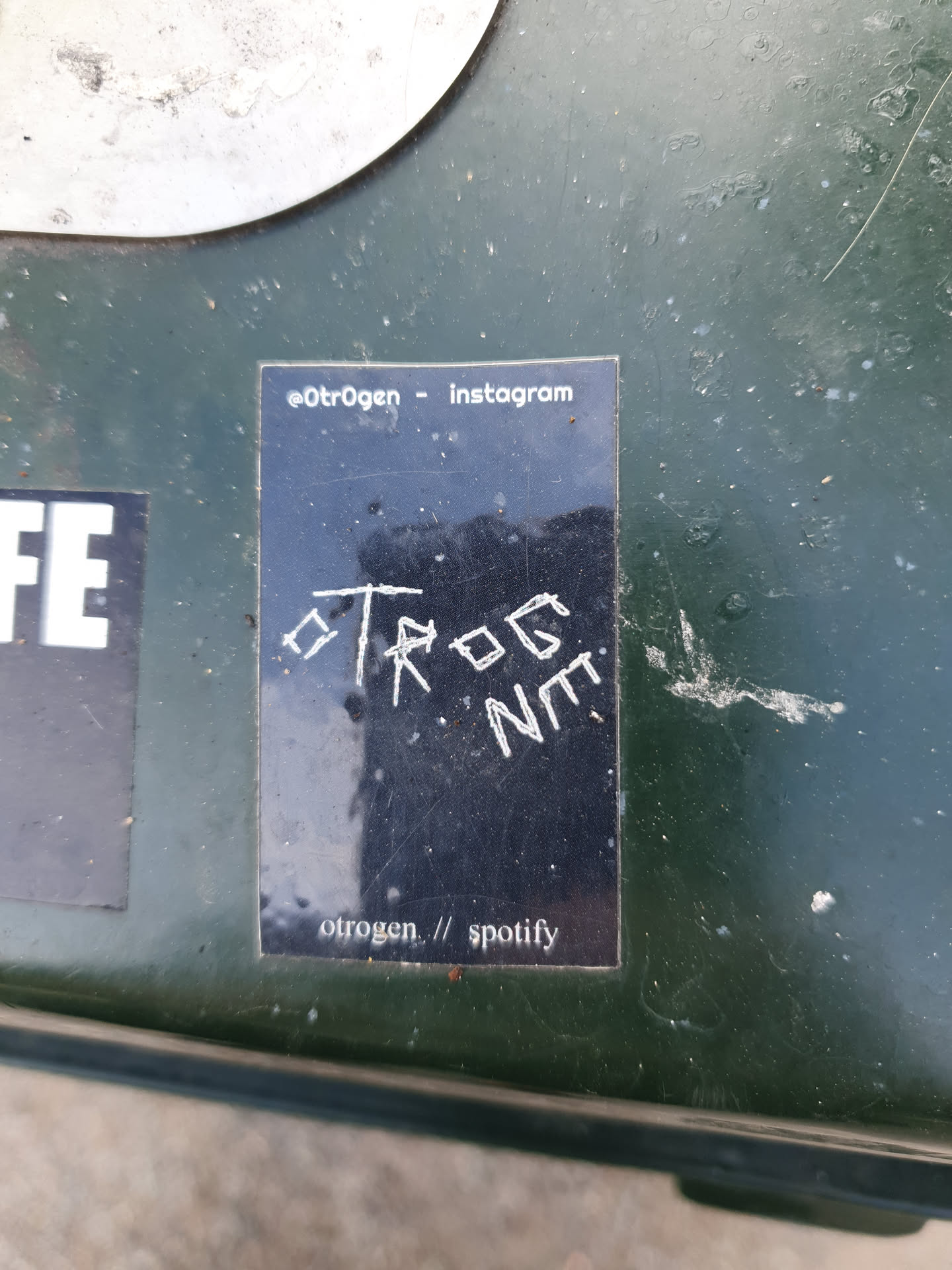 A black sticker with white text. The sticker has the artist's name oTrOgEn and social media links. The text on the sticker is hand-written and slightly messy.