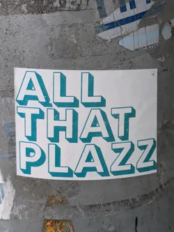 A teal and white sticker with the text "All That Plazz" in a stylized 3D font. The sticker is affixed to a weathered gray surface.
