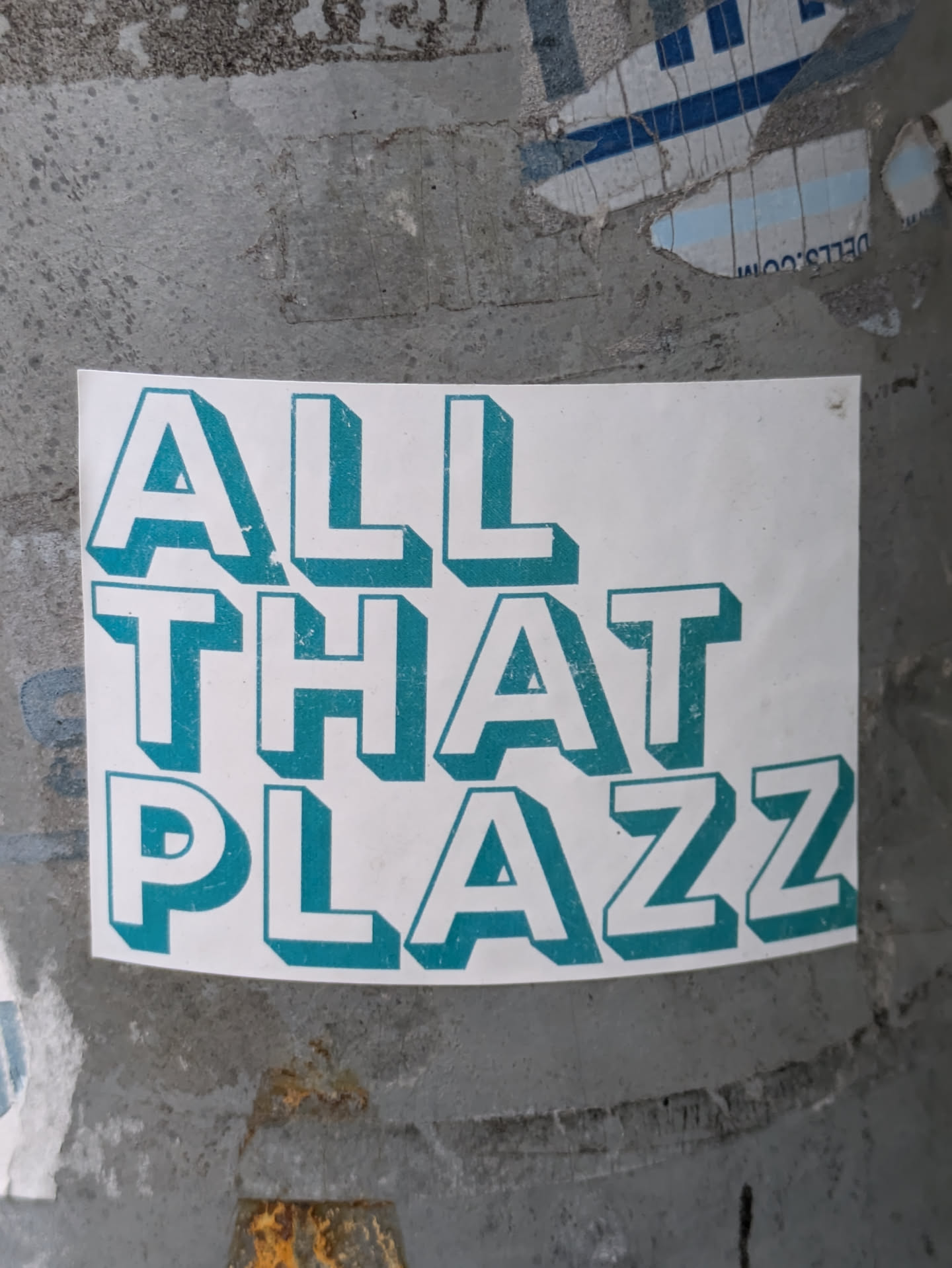 A teal and white sticker with the text "All That Plazz" in a stylized 3D font. The sticker is affixed to a weathered gray surface.