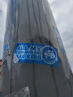 A blue sticker with a cartoon character and the text "All my friends are bad kids". The sticker is affixed to a gray pole outdoors.