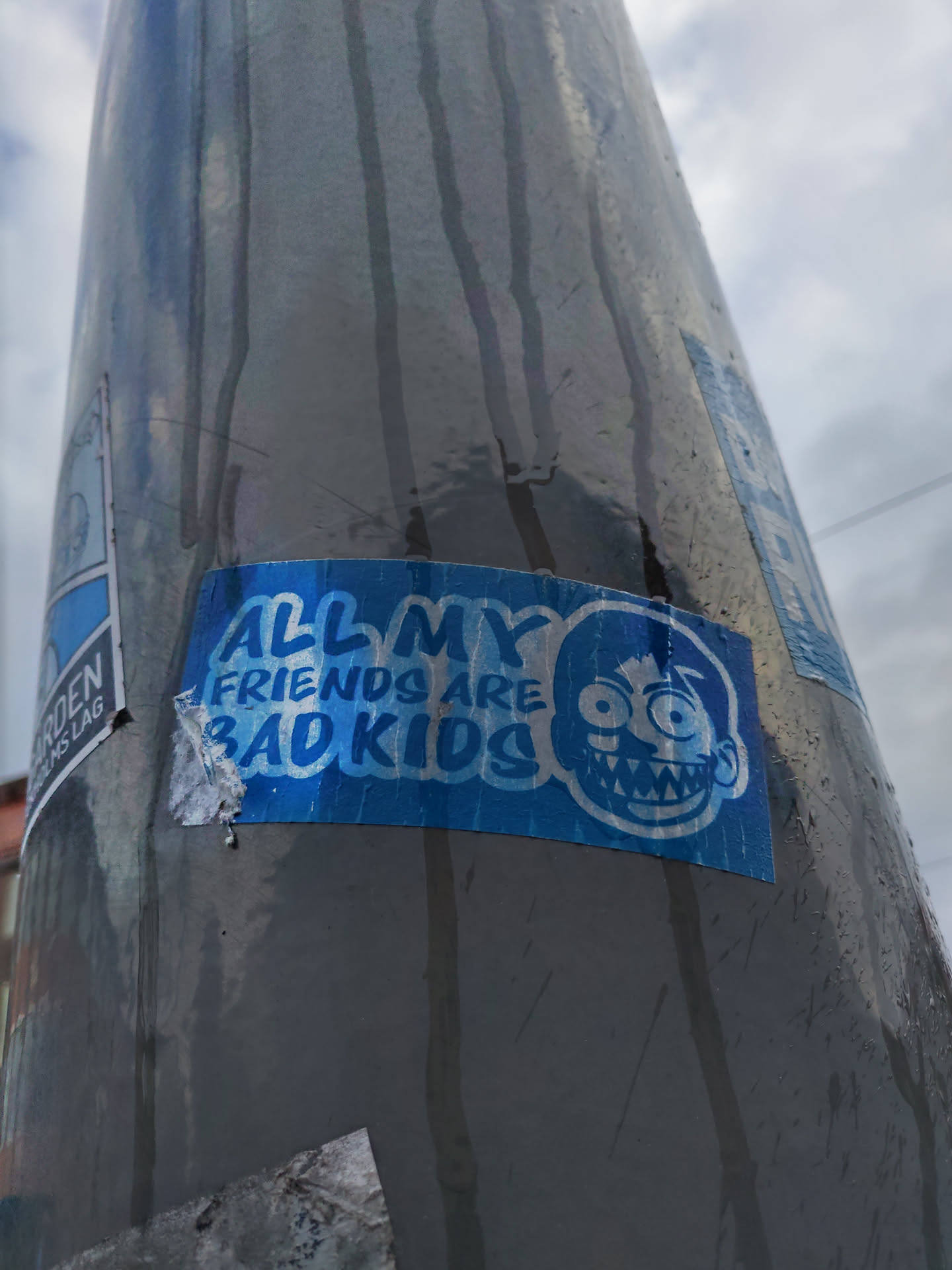 A blue sticker with a cartoon character and the text "All my friends are bad kids". The sticker is affixed to a gray pole outdoors.
