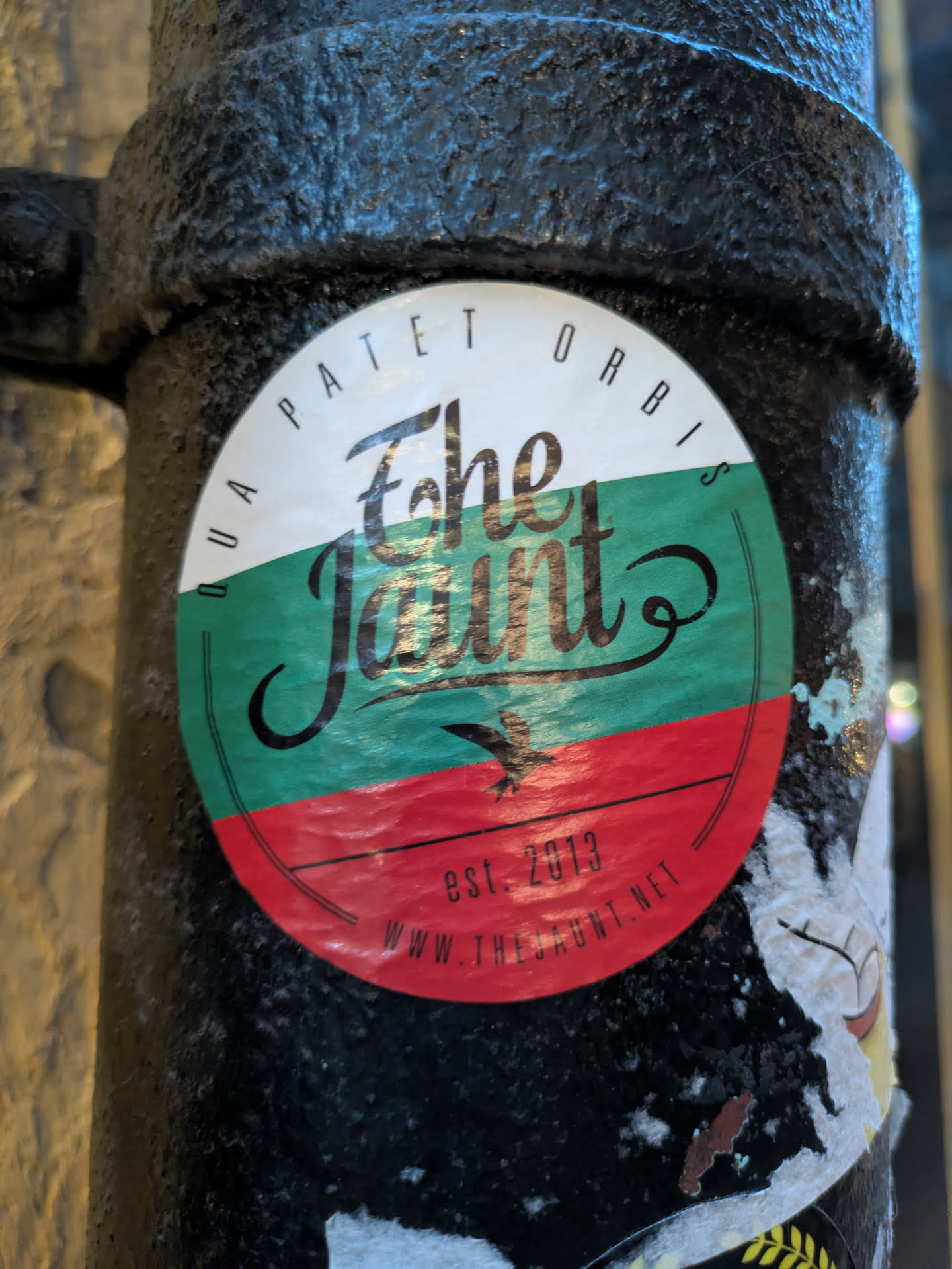 The sticker is circular and displays the national colors of Bulgaria: white, green, and red, arranged in horizontal stripes. The word 'The Jaunt' is written in a stylish, calligraphic font across the green and red stripes. Above the text, in a curved line, is the Latin phrase 'DUA PATET ORBIS'. Below the text, on the red stripe, is a small graphic of a bird in flight, and the text 'est. 2013' followed by 'WWW.THEJAUNT.NET'. The sticker appears to be slightly worn and attached to a dark, textured surface.
