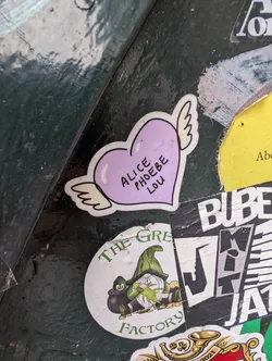 A purple heart-shaped sticker with small white wings attached to it. The text 'ALICE PHOEBE LOU' is written in black inside the heart. The sticker is placed among others.
