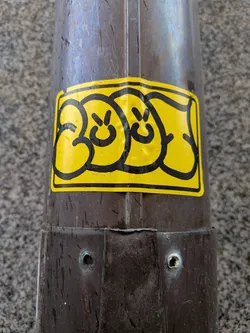 The sticker is a rectangular shape with rounded corners, featuring a vibrant yellow background. It displays three stylized, cartoonish faces or characters, each outlined with thick, bold black lines. Each face consists of two small black dots for eyes and a simple curved line for a mouth, conveying a generally smiley or neutral expression. The design has a distinct bubbly, graffiti-like aesthetic. The sticker appears slightly wrinkled and is covered with numerous small water droplets, suggesting it is outdoors and exposed to the elements. It is affixed to a dark, metallic pole.
