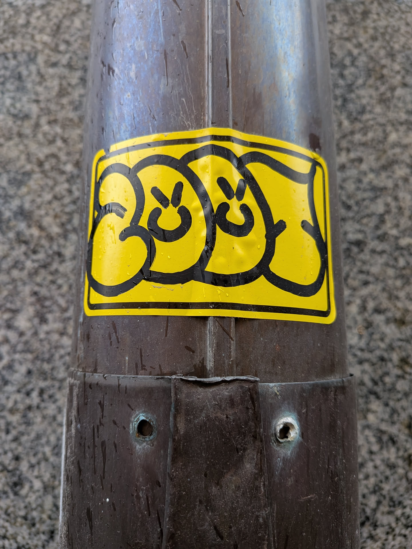 The sticker is a rectangular shape with rounded corners, featuring a vibrant yellow background. It displays three stylized, cartoonish faces or characters, each outlined with thick, bold black lines. Each face consists of two small black dots for eyes and a simple curved line for a mouth, conveying a generally smiley or neutral expression. The design has a distinct bubbly, graffiti-like aesthetic. The sticker appears slightly wrinkled and is covered with numerous small water droplets, suggesting it is outdoors and exposed to the elements. It is affixed to a dark, metallic pole.