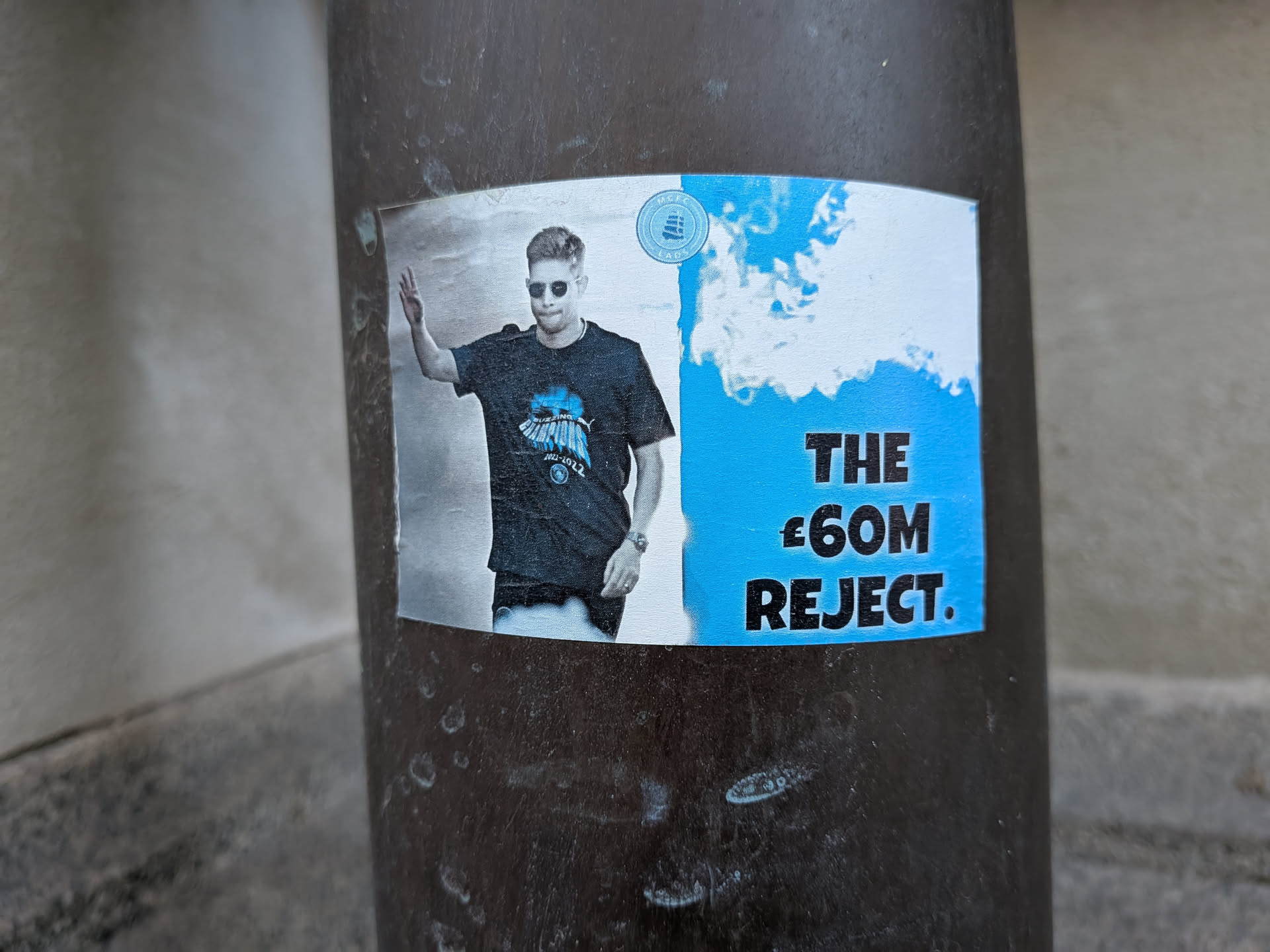 The sticker features a man wearing sunglasses and a t-shirt with a graphic on it. He is waving his right hand. The t-shirt has text and a graphic which appears to be a stylized depiction of something in blue. To the right of the man, on a bright blue background with white cloud-like shapes, are the words 'THE £60M REJECT.' in large black capital letters. There is also a circular emblem with blue and white elements visible above the man's head.