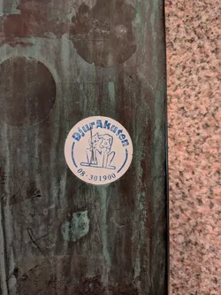 A circular white sticker with blue text and illustration, adhered to a dark, textured surface. The central illustration depicts two stylized animals, one resembling a cat and the other a dog, standing side-by-side. The text 'DjurAkuten' is curved along the top edge of the circle, and a phone number '08.301900' is curved along the bottom edge. The sticker shows signs of wear, including a vertical crease or tear through the center of the animal illustration and some minor scuffs.