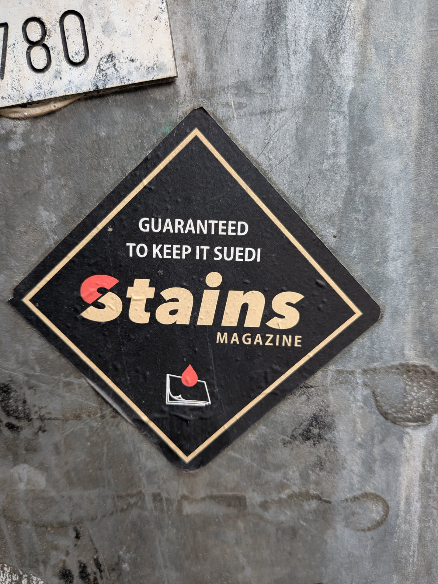 The sticker is diamond-shaped with a black background and a thin gold border. At the top, white text reads "GUARANTEED TO KEEP IT SUEDI". The prominent central element is the word "Stains" in large, stylized beige lettering, with the 'S' at the beginning featuring a red segment. Below "Stains" is the word "MAGAZINE" in smaller white capital letters. In the bottom right corner, there is a small graphic of a red droplet on top of stacked white papers, outlined in black.