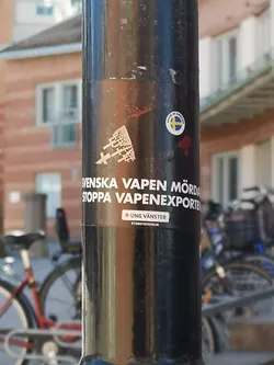 The central sticker is a dark, rectangular decal with white text and graphics, affixed to a black metal pole. At the top left, there's a white abstract graphic resembling fragmented 'X' shapes or a deconstructed weapon. Below this, the main message is prominently displayed in white, sans-serif capital letters: 'SVENSKA VAPEN MÖRDAR STOPPA VAPENEXPORTEN' (Swedish weapons kill, stop weapon exports). Further down, a smaller white rectangular box contains a red star icon followed by 'UNG VÄNSTER' (Young Left), and below that, 'STORSTOCKHOLM' (Greater Stockholm). Slightly above and to the right of the main sticker, there's a smaller circular sticker with a white border, featuring the text 'MADE IN SWEDEN' encircling a depiction of the Swedish flag. The sticker shows some minor wear and tear, consistent with being outdoors.