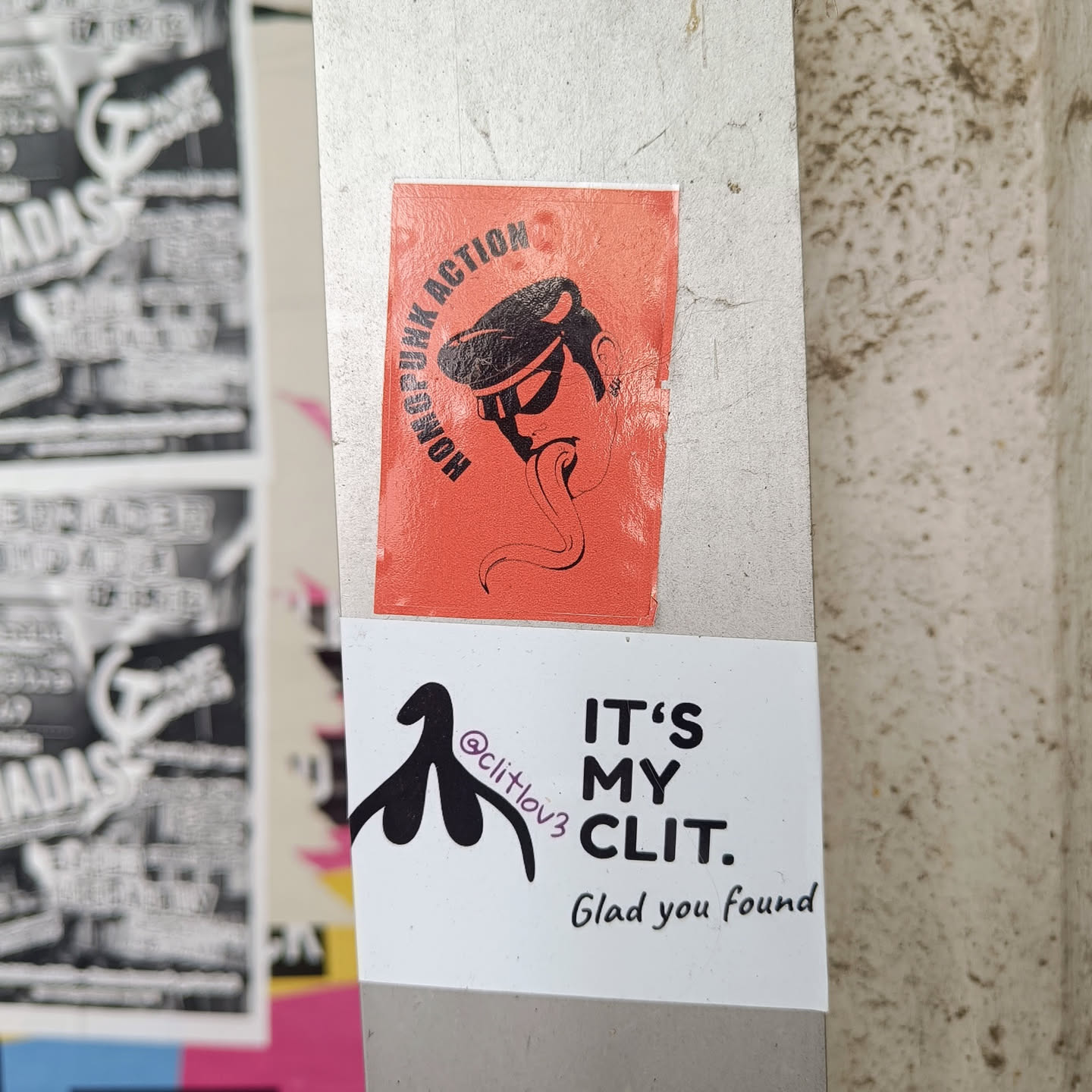 Two stickers on a pole. The top one is orange with a black illustration of a head with a cap and a long tongue. The text on the orange sticker says "HORSEF*NK ACTION". The bottom sticker is white with a black illustration of a clitoris and the text "IT'S MY CLIT. Glad you found". There is also a social media handle on the white sticker: @clitlov3
